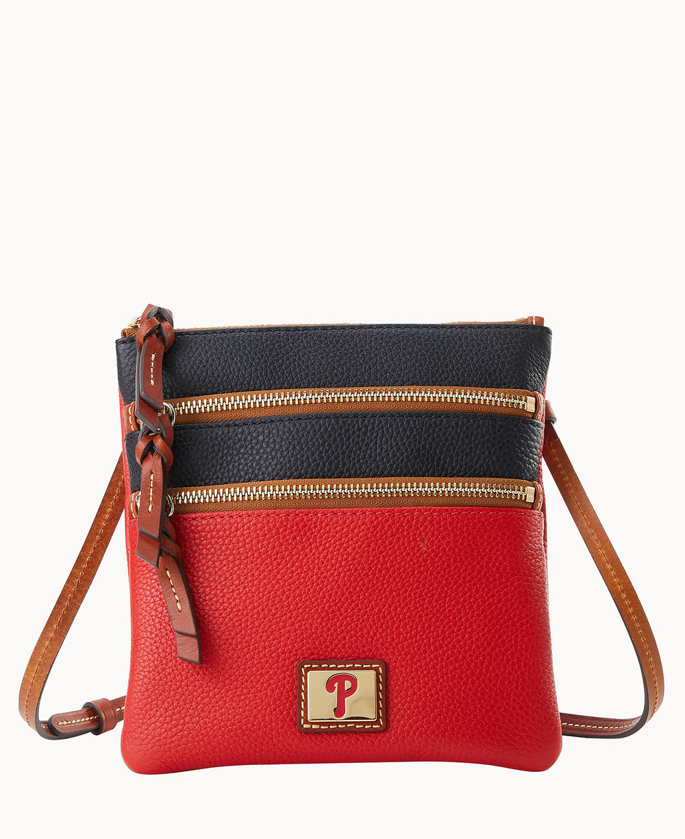 A small leather crossbody bag with a red bottom half, black top half, two gold zippers, a tan adjustable strap, and a square patch with a P logo on the front.