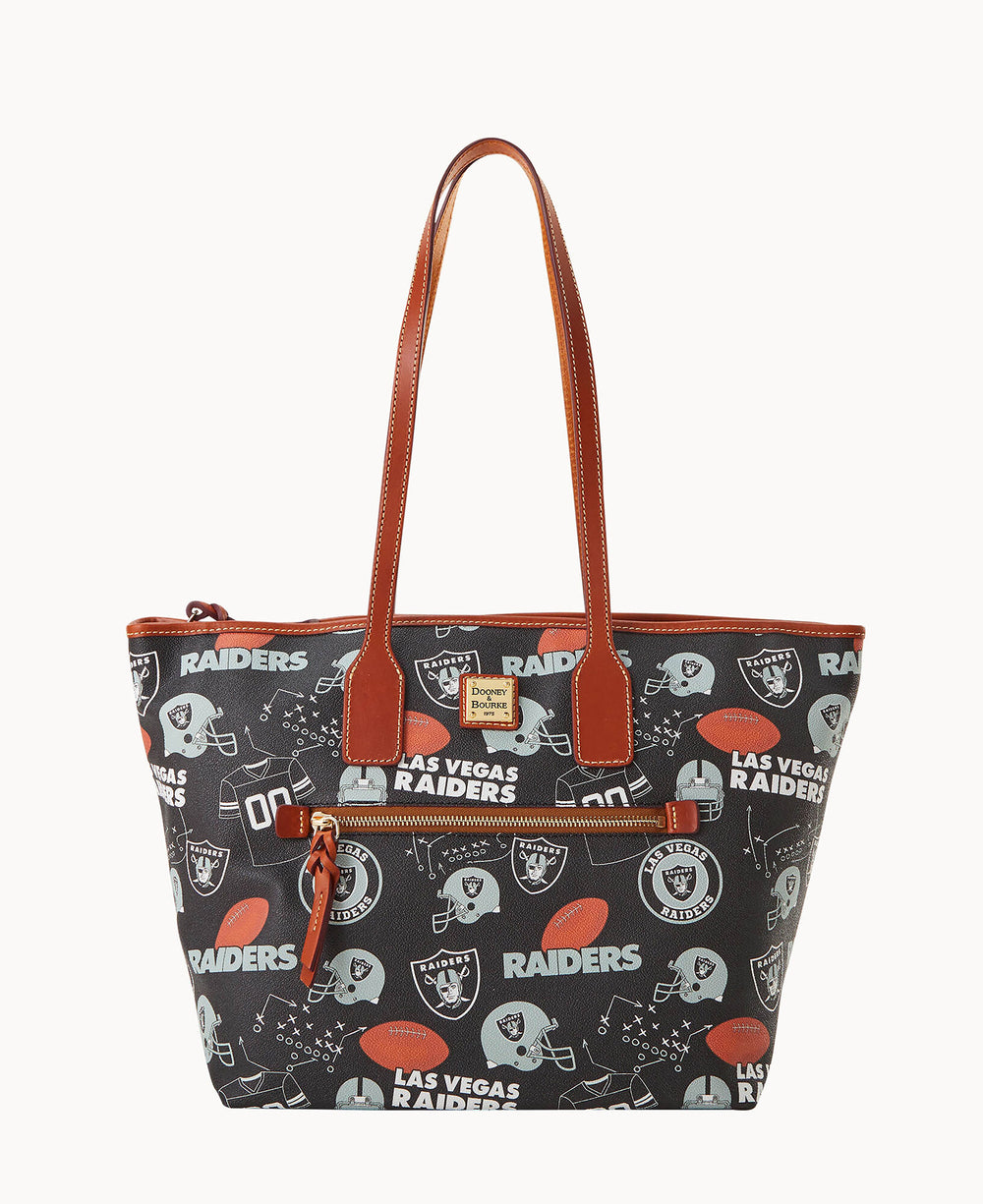 A black tote bag with brown leather handles and trim, featuring a Las Vegas Raiders football team print, including logos, helmets, and the team name. The bag has a front zip pocket and a zipper closure on top.