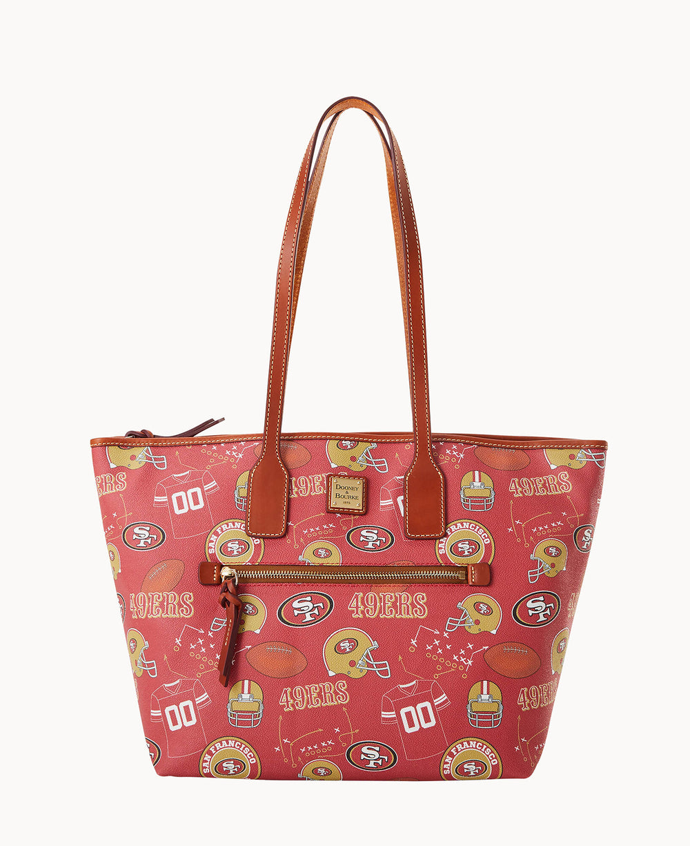 A red tote bag with brown handles featuring San Francisco 49ers logos, football helmets, and “49ERS†text in a repeating pattern, with a front zipper pocket and gold-tone brand plate.