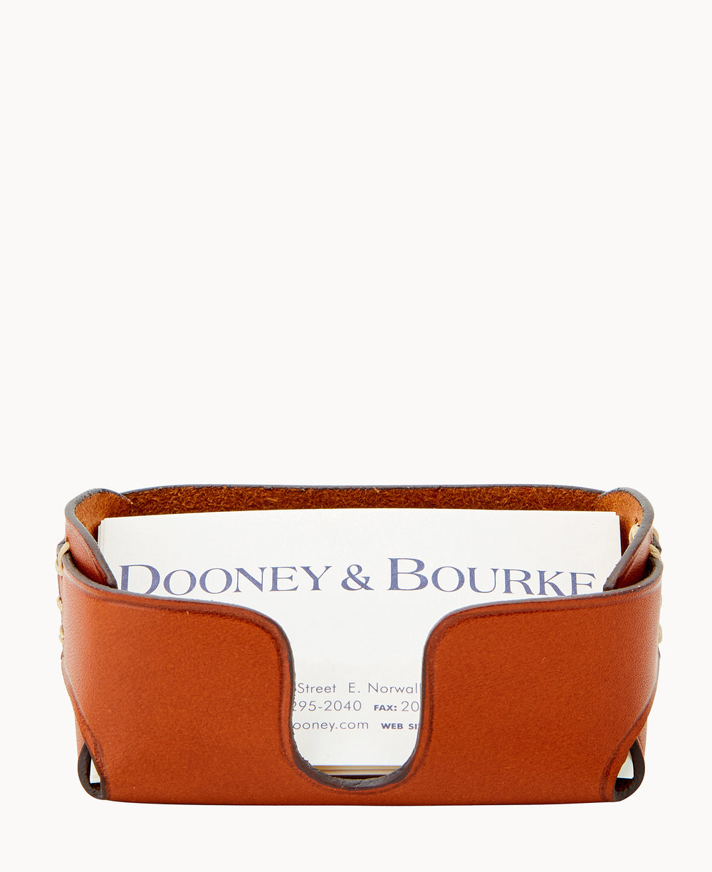 A tan leather business card holder with a curved front edge, displaying a Dooney & Bourke business card standing upright inside it. The background is plain white.
