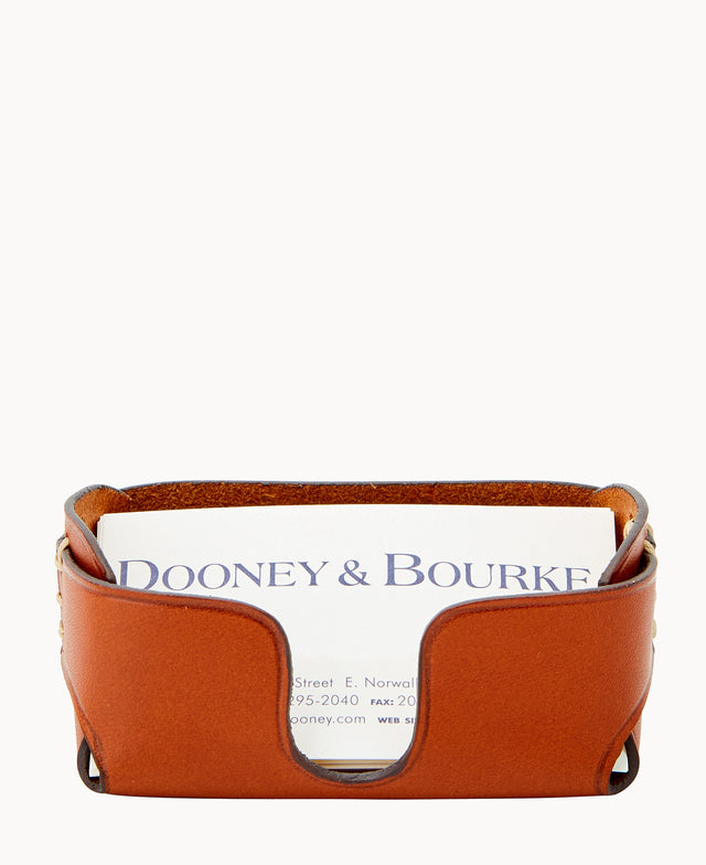 A tan leather business card holder with a curved front edge, displaying a Dooney & Bourke business card standing upright inside it. The background is plain white.