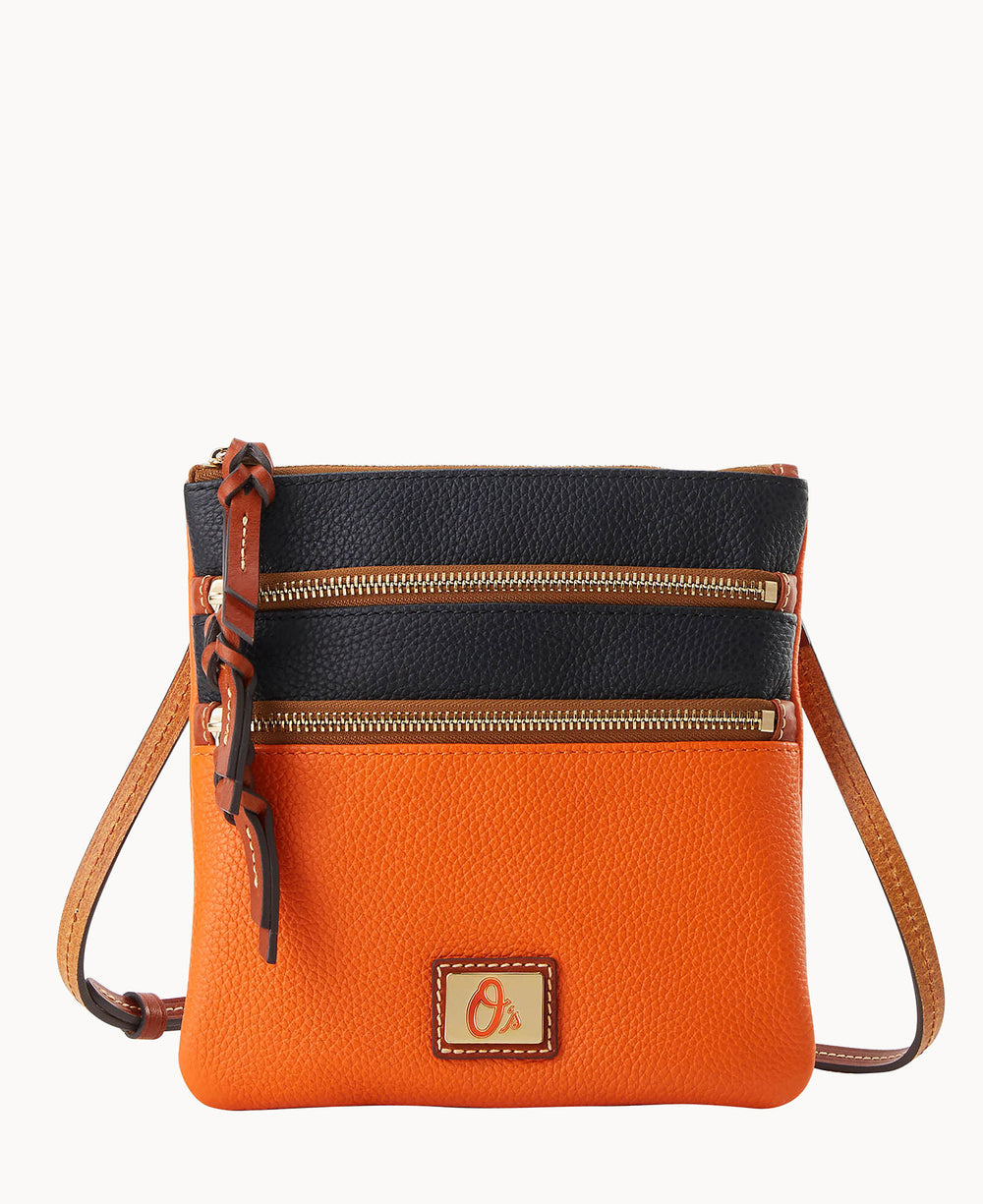 A small crossbody bag with an orange bottom half, black top half, two front zippered pockets, a tan adjustable strap, and a decorative leather tassel. The bag features a rectangular logo patch on the front.