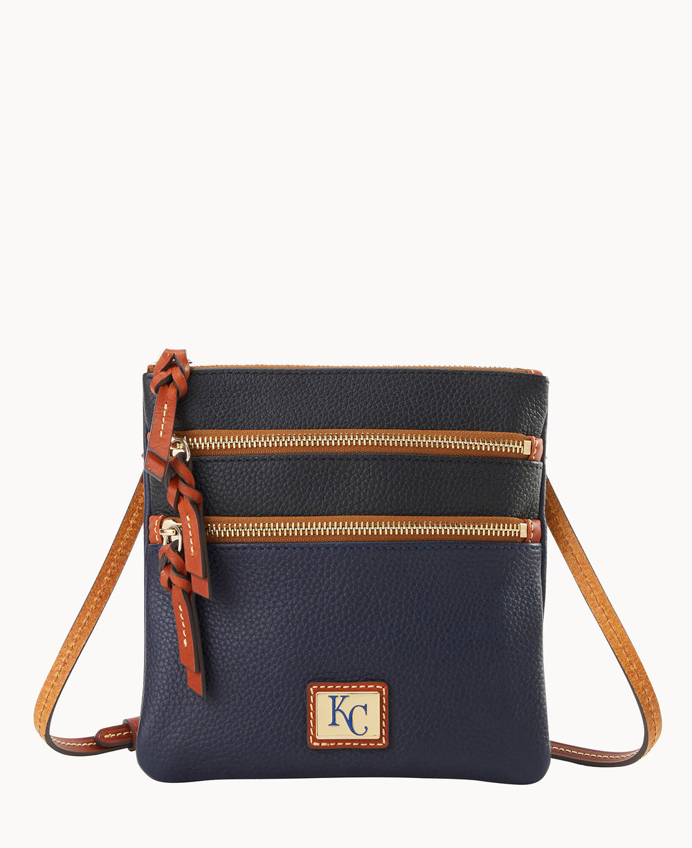A navy blue crossbody bag with brown leather trim, two front zipper pockets with tassel pulls, and a tan shoulder strap. The front features a rectangular KC logo patch.