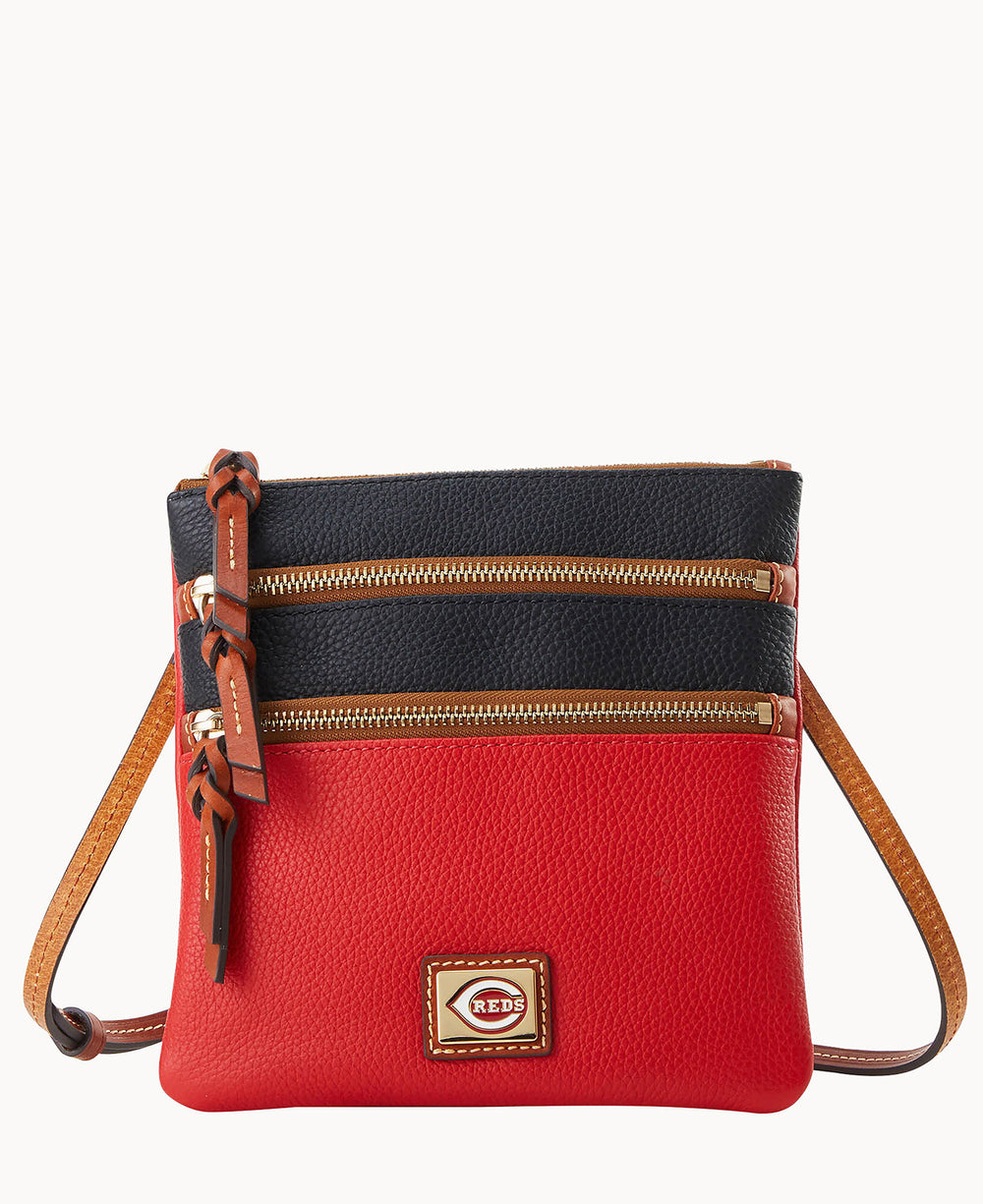 A small crossbody bag with a red bottom half, black top half, two front zippered pockets with brown leather pulls, a brown adjustable strap, and a small rectangular logo patch on the front.