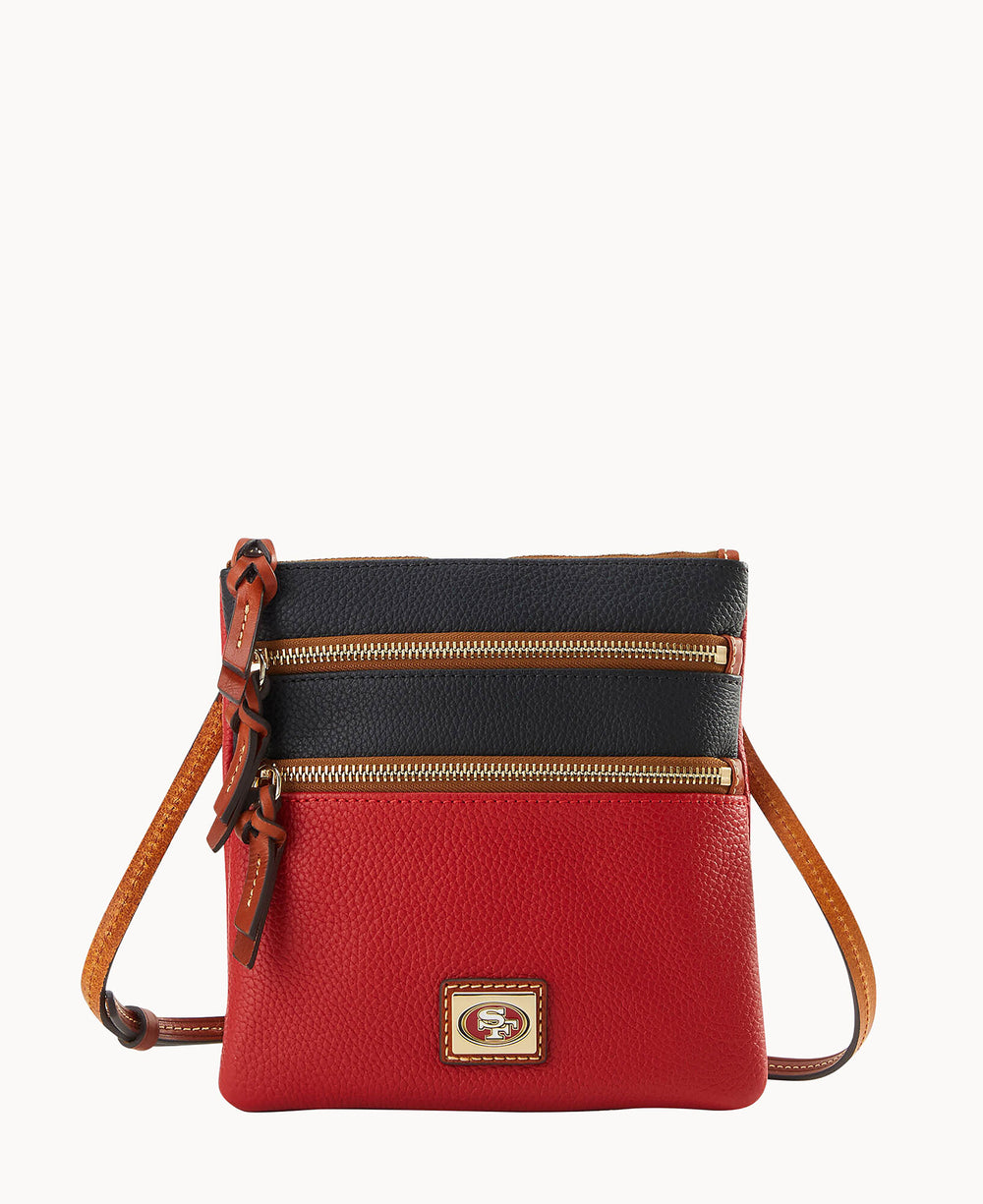 A small crossbody bag with a red bottom, black top, tan strap, and two front zippered pockets. The bag features a gold and brown logo plate on the front.