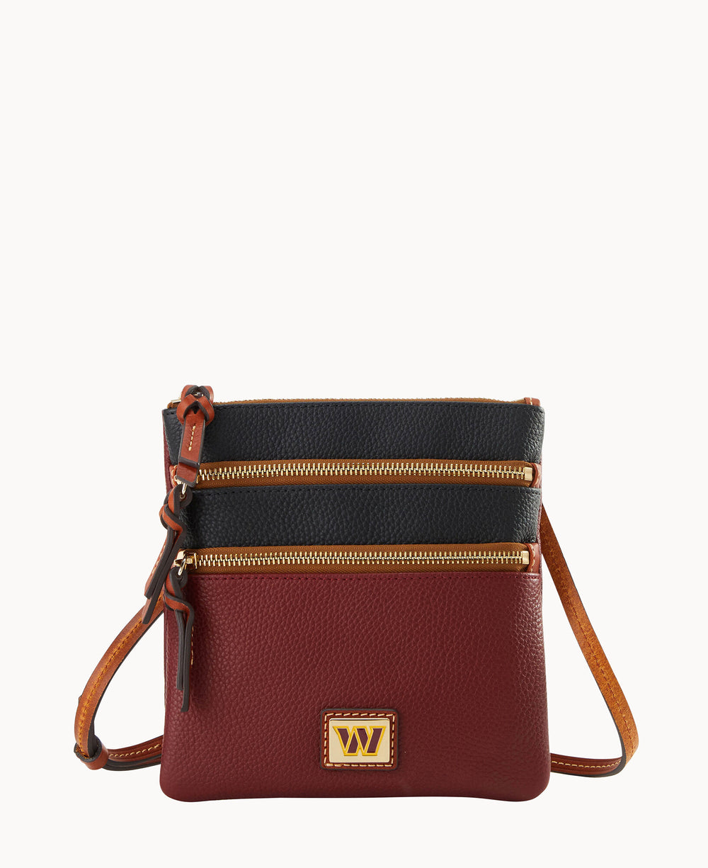 A small crossbody bag with a black top section, burgundy bottom section, two gold zippered pockets, a tan adjustable strap, and a gold and brown rectangular logo plate on the front.