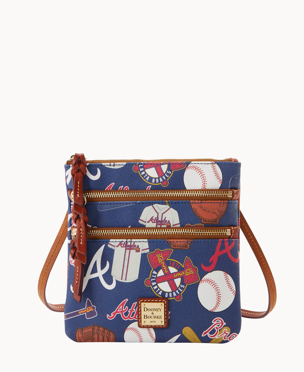 A small crossbody bag with brown leather straps and trim, decorated with images of baseballs, gloves, jerseys, and Atlanta Braves logos on a blue background. The bag has two front zippered pockets.