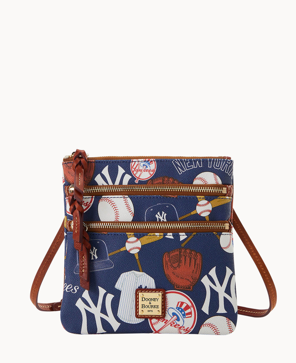 A small crossbody bag with a blue background featuring New York Yankees logos, baseballs, bats, gloves, and caps. The bag has two front zippered pockets and a brown leather strap and accents.