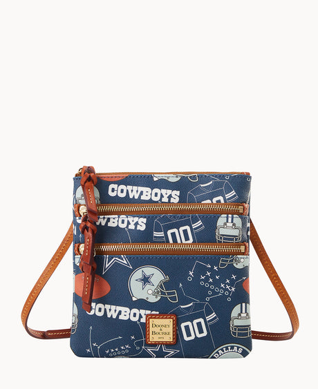 A small crossbody bag with brown leather straps and trim, featuring a blue pattern with Dallas Cowboys logos, football helmets, footballs, and the word COWBOYS in white. The bag has two front zippered pockets.