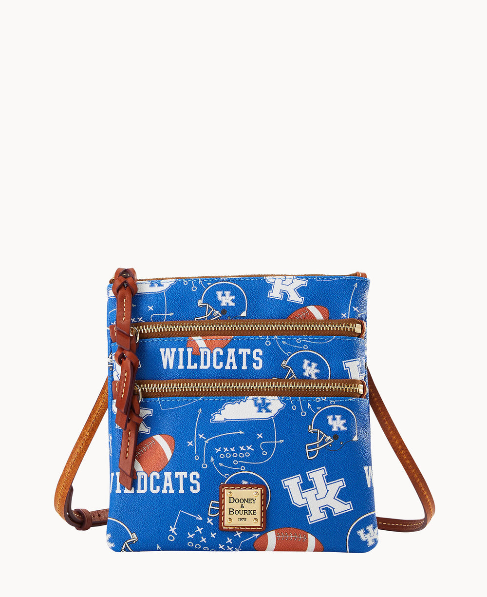 A small blue crossbody bag with brown leather accents, featuring University of Kentucky Wildcats logos, footballs, and play diagrams. The bag has two front zippered pockets and a Dooney & Bourke label.