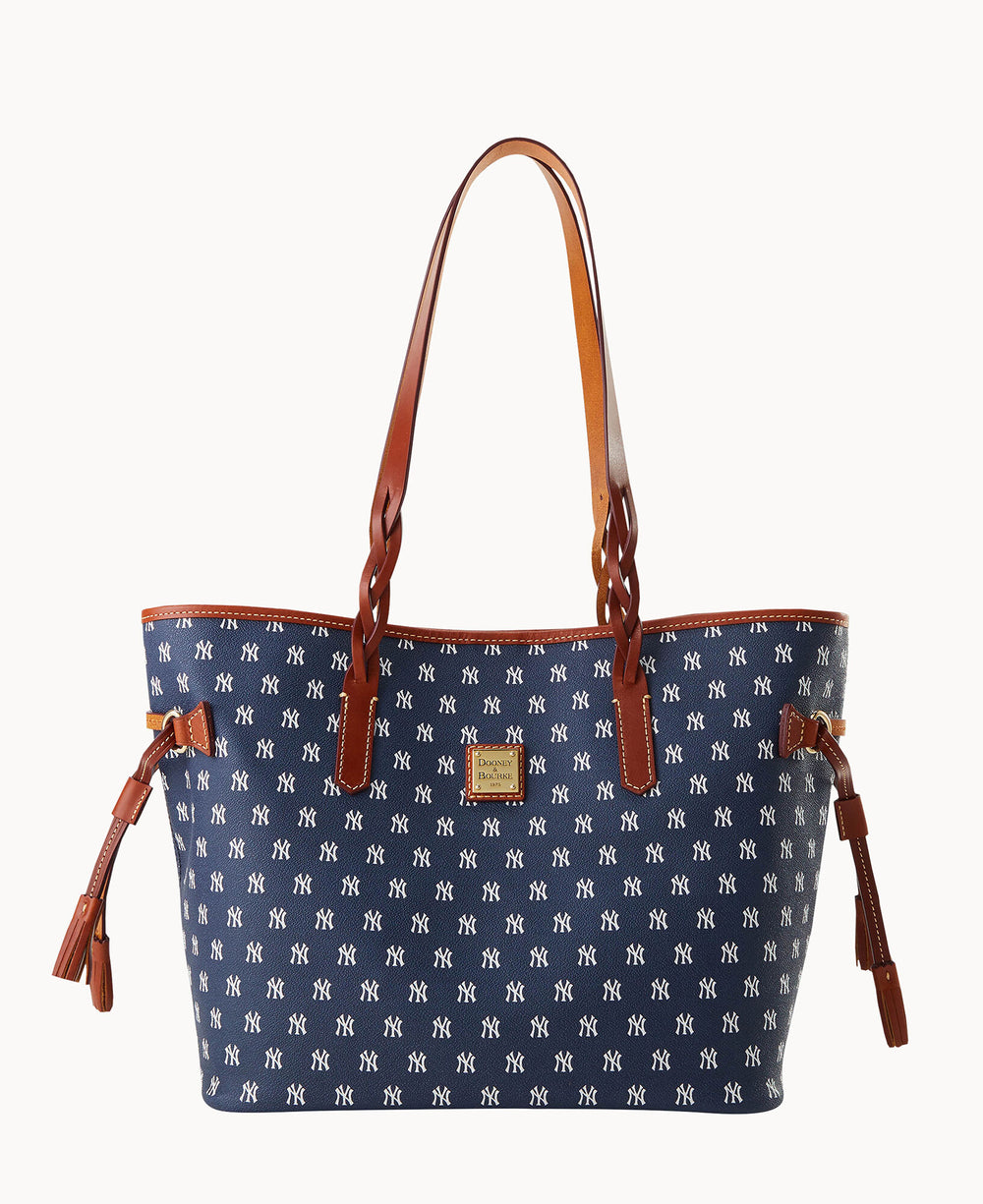 A navy blue tote bag with brown leather handles and trim, featuring an all-over white repeating NY pattern. The bag has a rectangular shape and tassel accents on the sides.