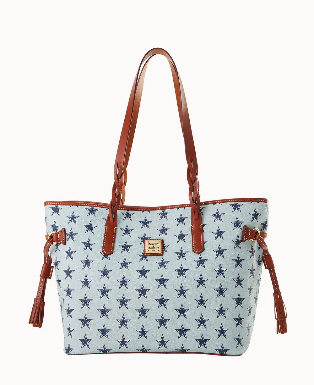 A light blue tote bag with dark blue star patterns, brown leather handles, brown leather tassels on the sides, and a small rectangular logo patch on the front.