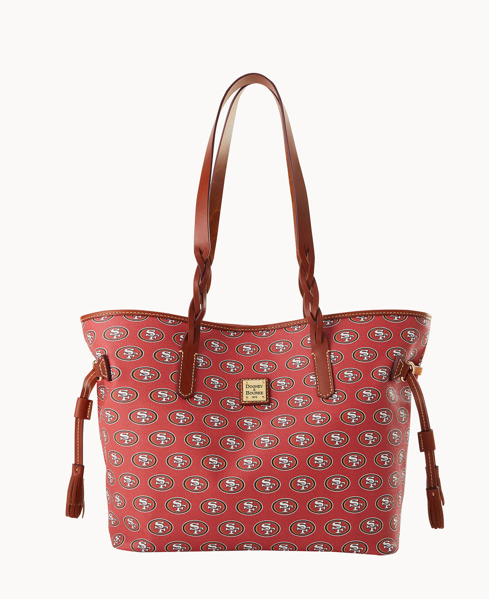 A red tote bag with brown handles features a repeating pattern of the San Francisco 49ers logo and a gold Dooney & Bourke label on the front.
