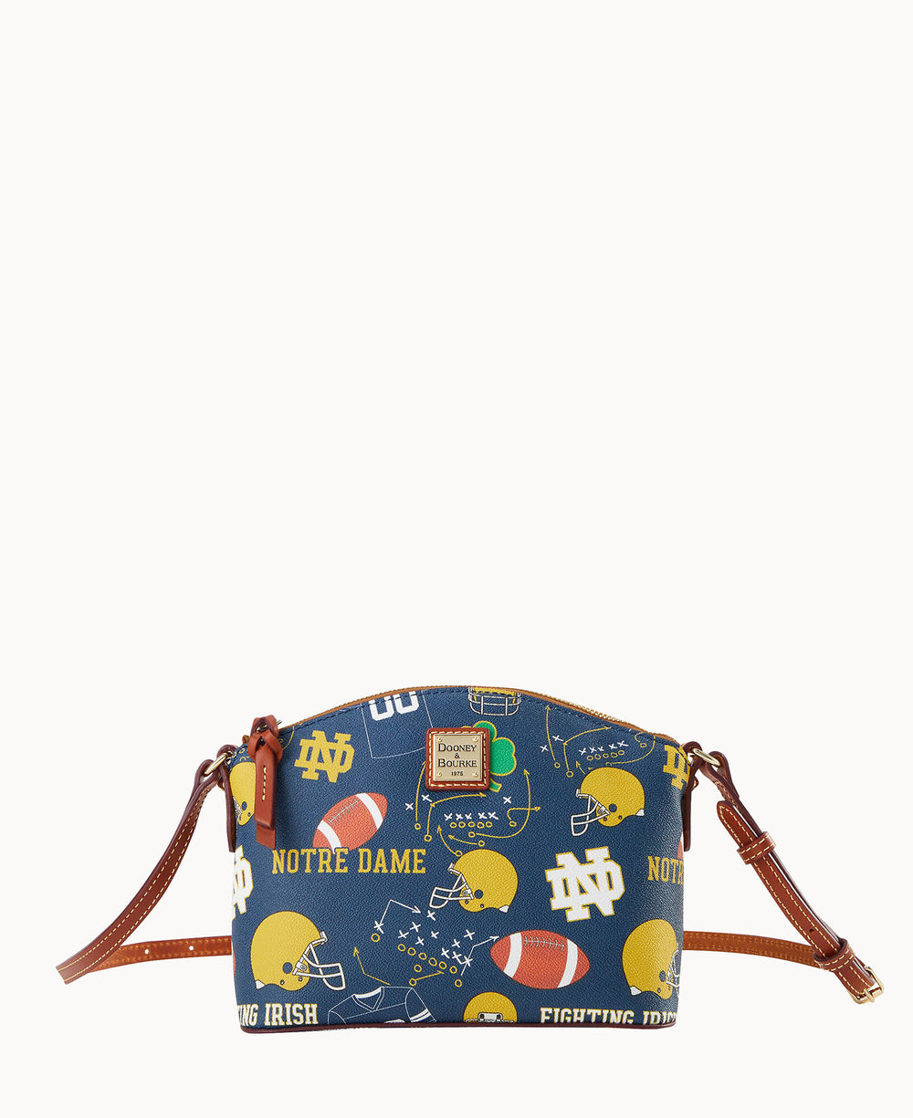 A blue Dooney & Bourke crossbody bag with brown leather straps, featuring a Notre Dame theme with yellow helmets, footballs, and Notre Dame logos and text in yellow and white.