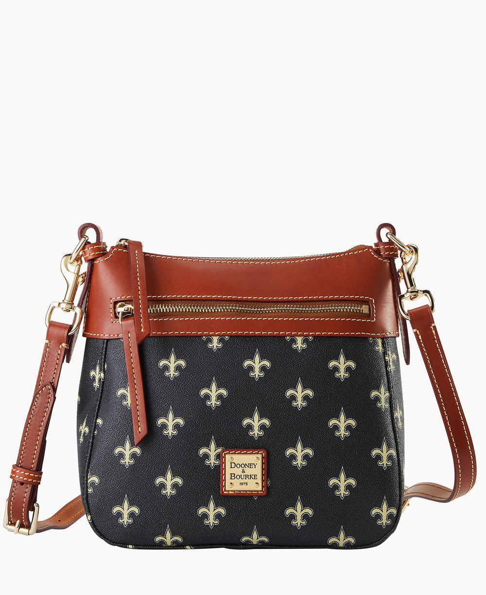 A black Dooney & Bourke crossbody bag with tan leather trim, gold hardware, front zipper pocket, and a repeating fleur-de-lis pattern. The logo patch is centered at the bottom front of the bag.