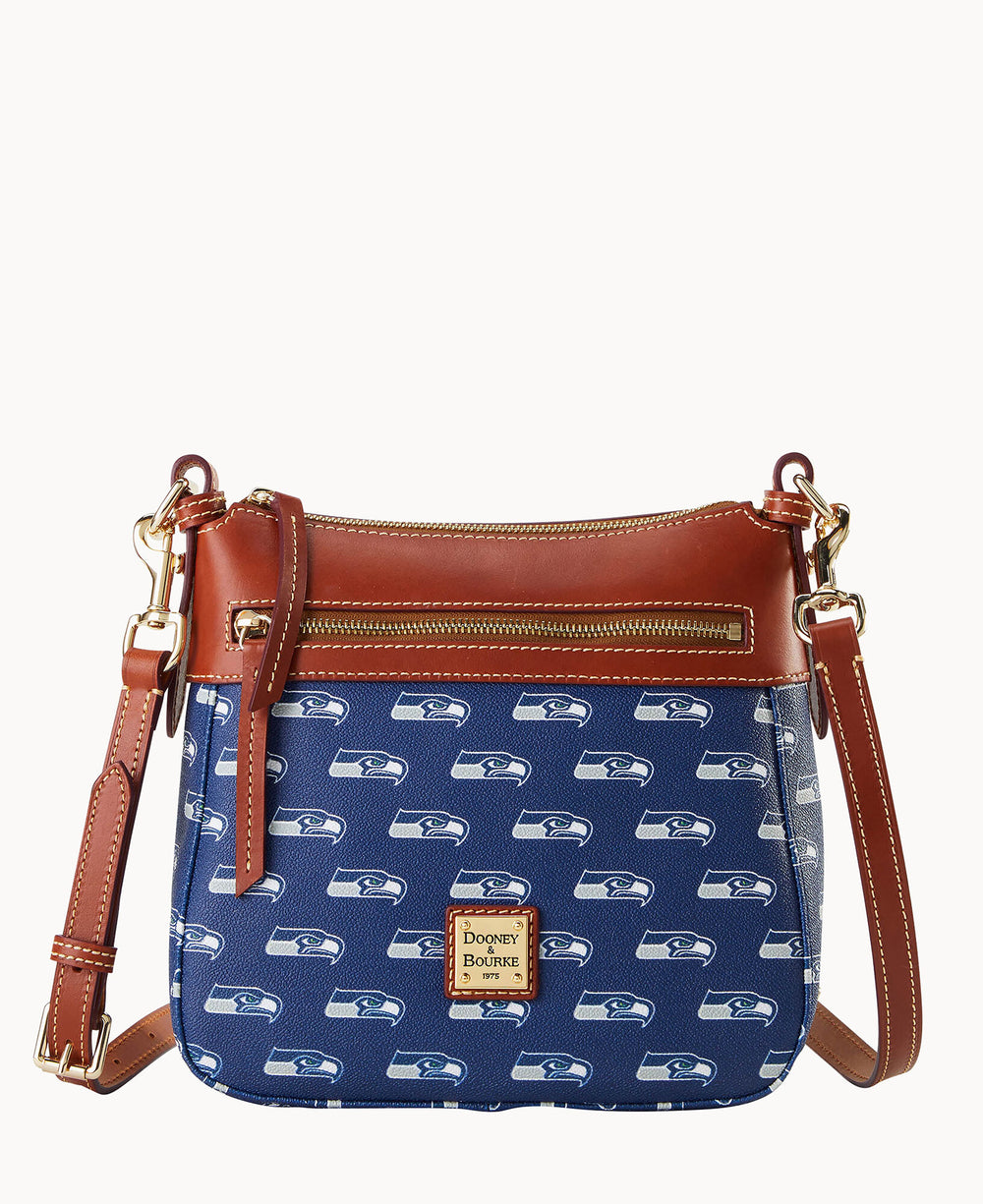 A blue crossbody bag with brown leather accents, a front zipper pocket, and a gold label. The bag features a repeating white Seattle Seahawks logo pattern and has an adjustable strap.