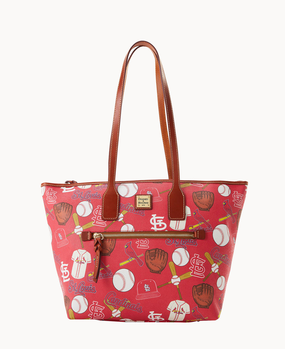 A red tote bag with brown handles, featuring St. Louis Cardinals baseball-themed graphics including balls, gloves, hats, and team logos. There is a front zip pocket and a small logo plate above it.