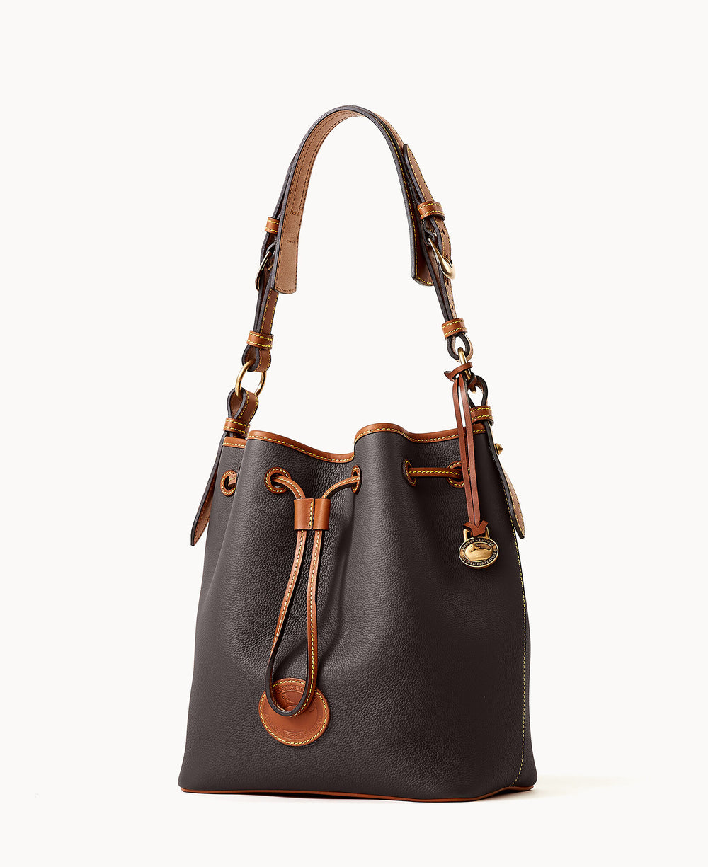 A brown leather bucket bag with tan leather trim, a drawstring closure, gold hardware, and a short shoulder strap. The bag has a structured shape and a decorative logo charm hanging on one side.