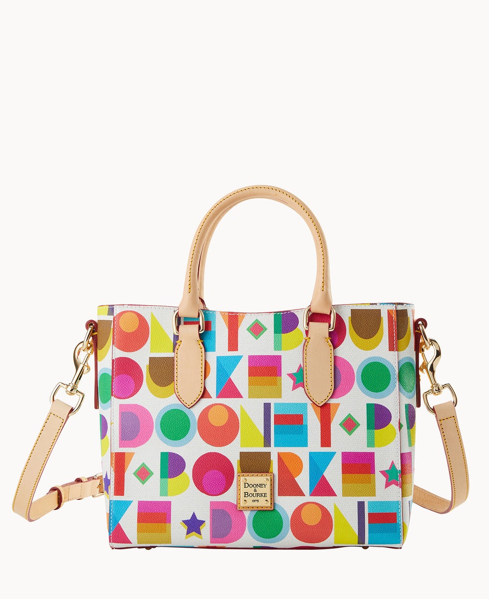 A handbag with tan leather handles and a detachable shoulder strap, featuring a colorful geometric pattern with circles, squares, triangles, stars, and letters on a white background.