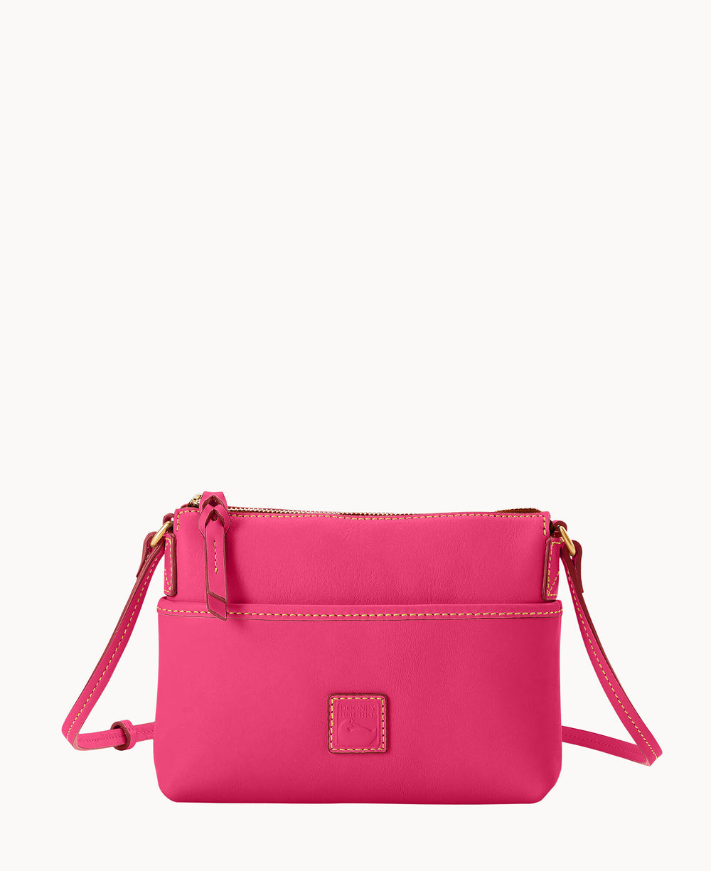 A small, bright pink crossbody purse with a zipper closure, gold hardware, and an adjustable strap. The bag features white stitching and a square stitched logo patch on the front.