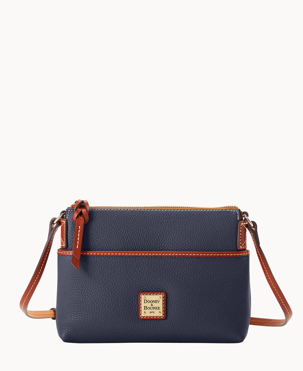 A small navy blue crossbody bag with brown leather trim, gold-tone hardware, and a front logo patch. The bag has a zippered top and an adjustable strap.