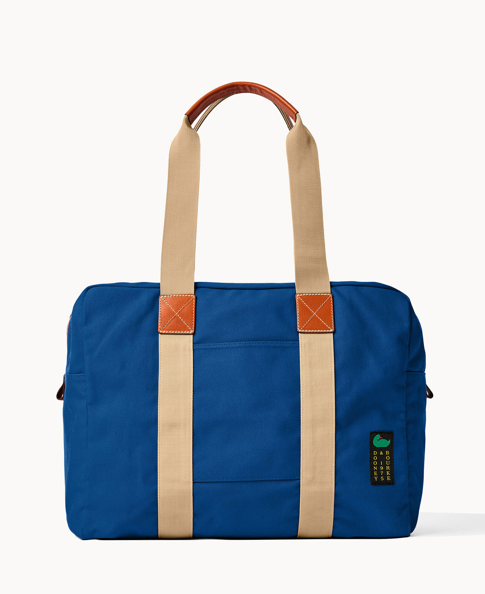 A blue canvas duffel bag with tan straps, brown leather accents, and a small green and black label on the front. The bag is photographed against a plain white background.