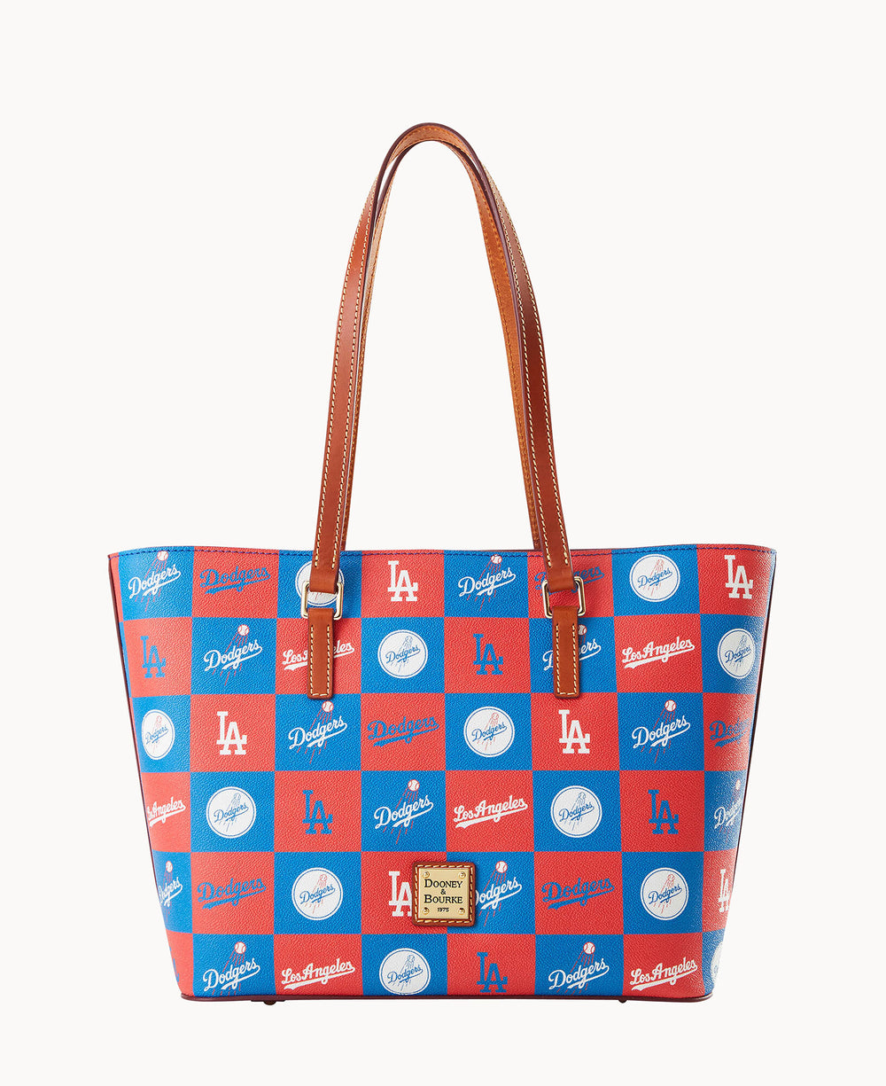 A tote bag with brown handles features a red and blue checkered pattern decorated with white LA Dodgers logos and text. A gold and brown Dooney & Bourke label is attached to the front.