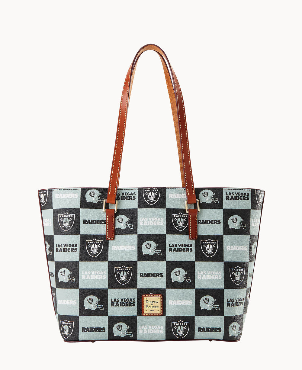 A tote bag with brown leather handles features a checkered pattern of Las Vegas Raiders logos and text in black, gray, and white. The Dooney & Bourke label is displayed on the front.