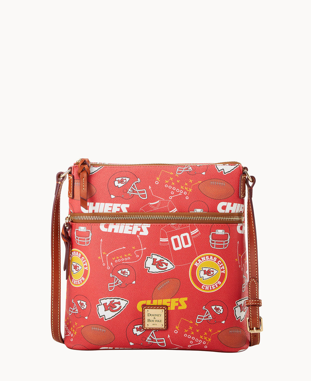 A red crossbody bag with Kansas City Chiefs logos, football helmets, and football graphics, featuring gold zippers and a brown adjustable strap. The bag has a front zip pocket and a small Dooney & Bourke label.