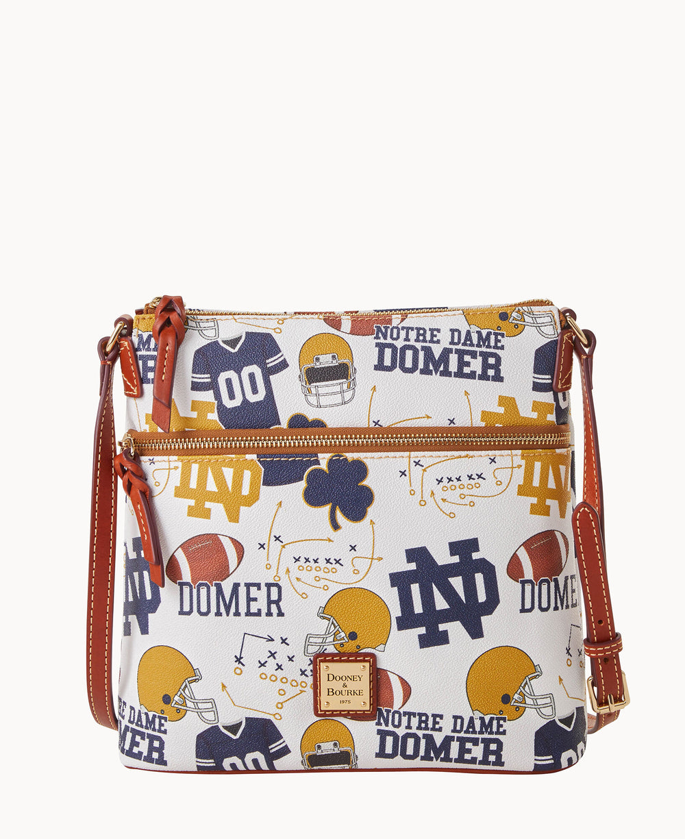 A white crossbody bag with brown straps featuring Notre Dame football-themed patterns, including helmets, jerseys, shamrocks, footballs, and Notre Dame Domer text in blue and gold.
