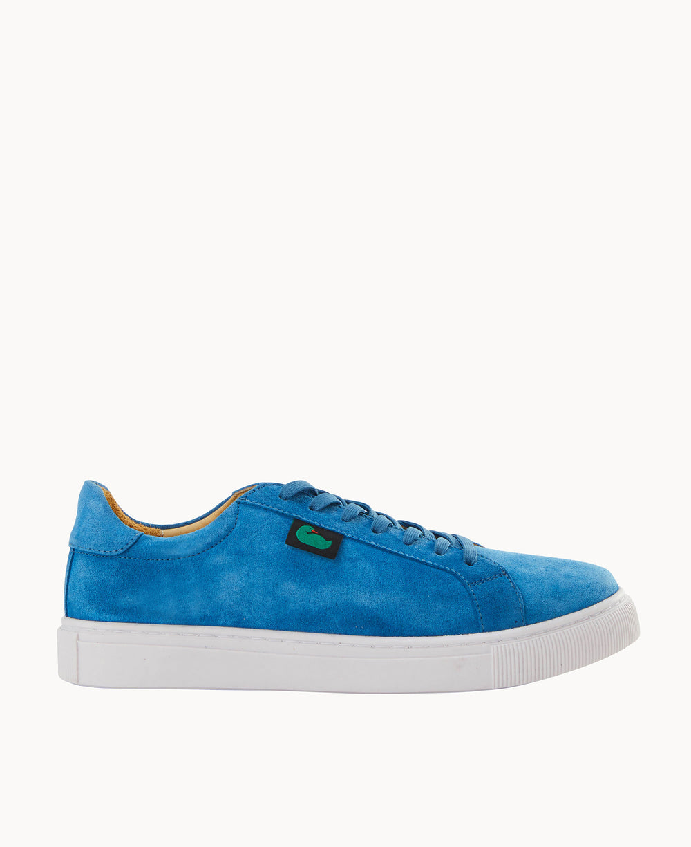 A bright blue suede sneaker with matching blue laces, a white rubber sole, and a small black rectangular tag with a green logo on the side, shown in a side view against a white background.