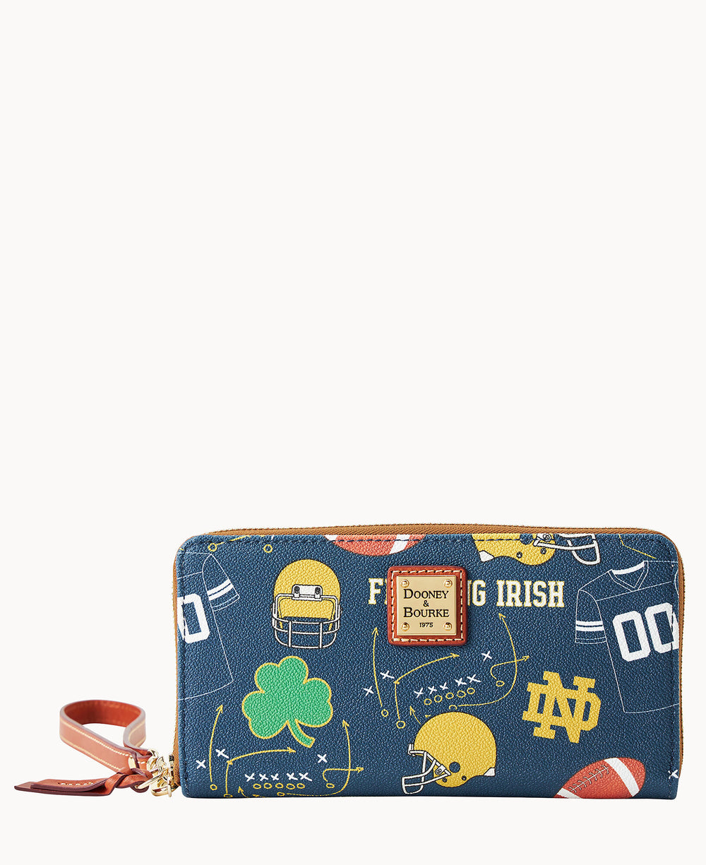 A blue Dooney & Bourke zippered wallet with football-themed Notre Dame graphics, including helmets, footballs, a shamrock, and the words â€œFighting Irish,â€ with a brown leather wrist strap.